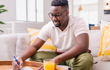 Person seated on a couch at home, filling out paperwork on a coffee table near a glass of orange juice.