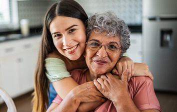 Person embraces an older relative from behind in a bright kitchen, both smiling warmly at the camera.