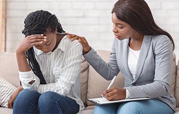 Woman receives emotional support from another woman while seated on a couch.