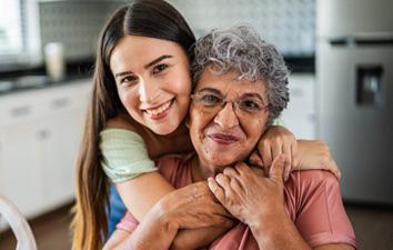 Person embraces an older relative from behind in a bright kitchen, both smiling warmly at the camera.