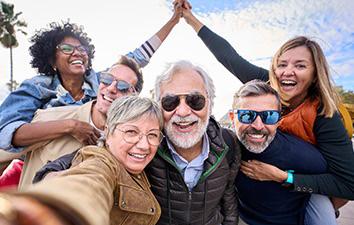 Group of five older adults smiling and laughing together, leaning in for a cheerful outdoor selfie.
